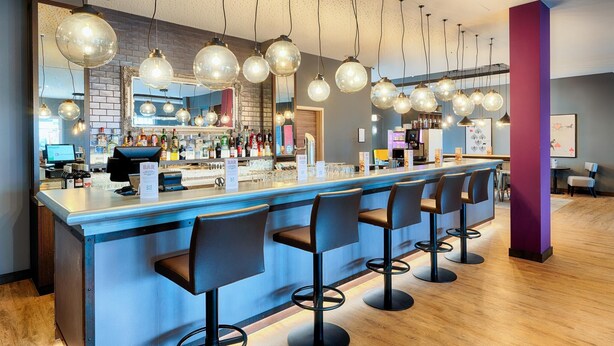 A bar area with a long counter, multiple stools, hanging spherical light fixtures, and a brick wall backdrop. - Premier Inn Lindau (Lindau)