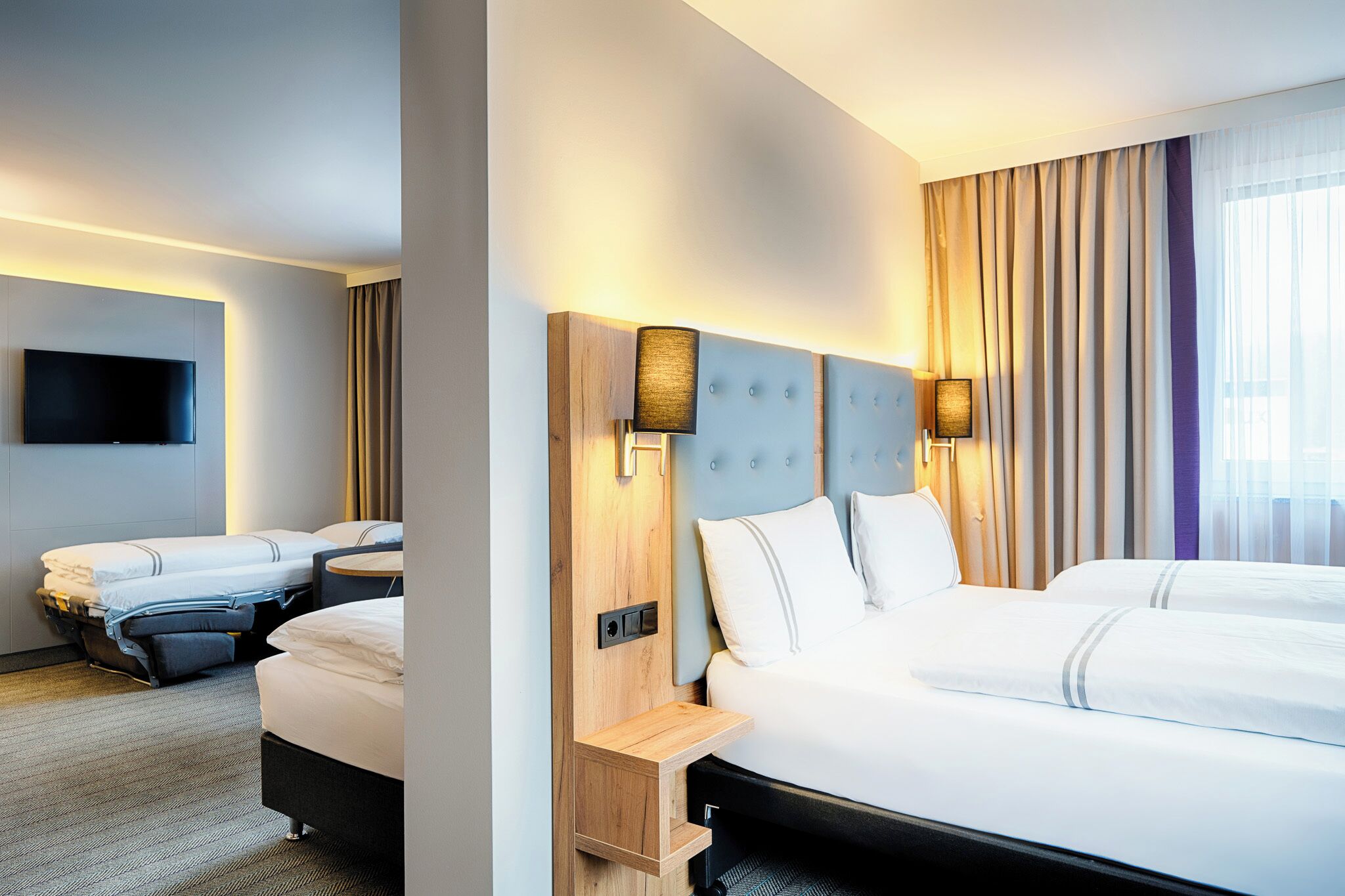Habitación de hotel con una cama grande, una cama más pequeña, un televisor montado en la pared y una ventana con cortinas.
