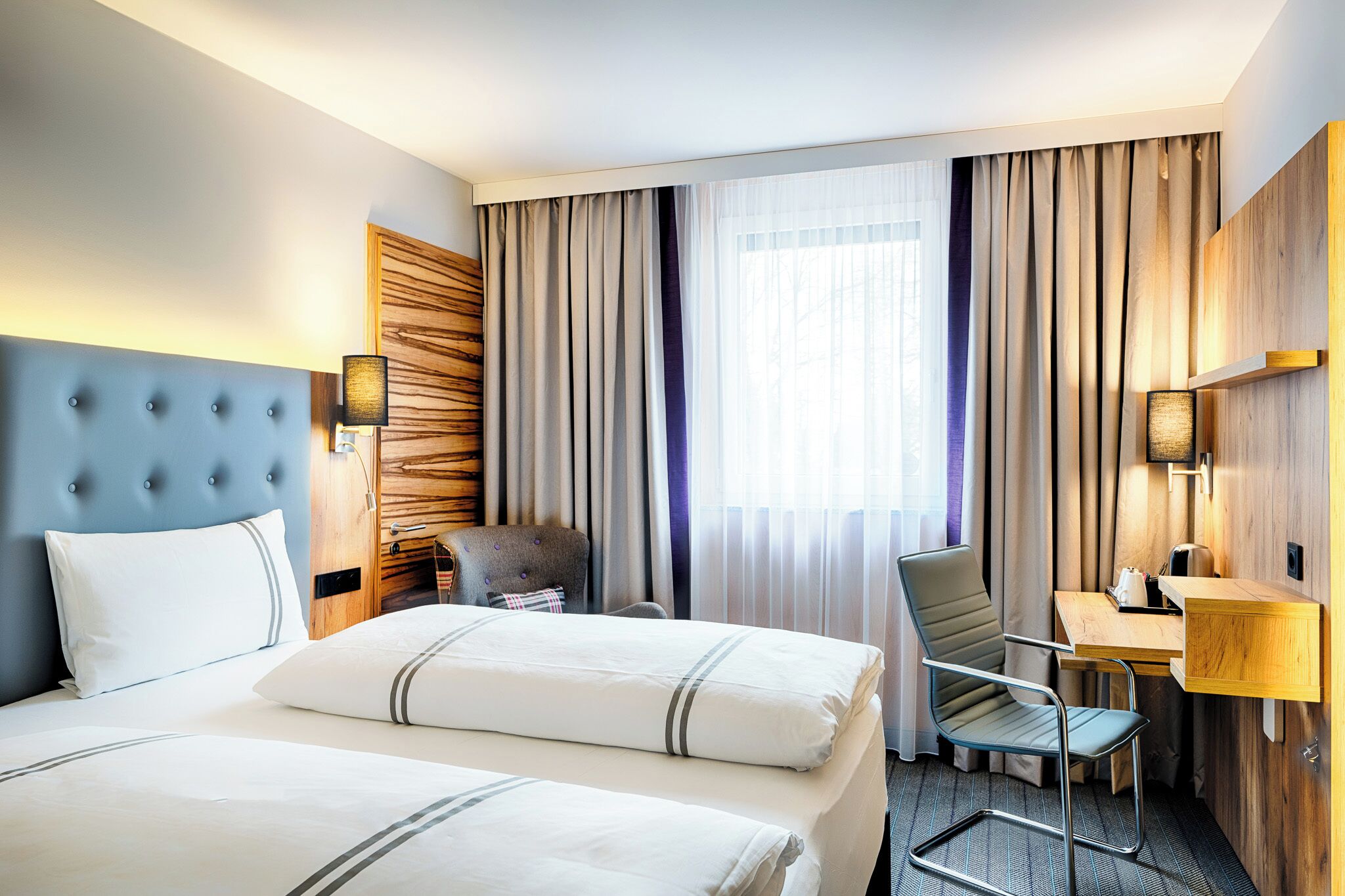 A hotel room with two beds, a desk, a chair, and a large window with curtains.