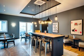 A modern bar area with a central island, bar stools, pendant lighting, and framed artwork on the walls. - Premier Inn Lindau (Lindau)
