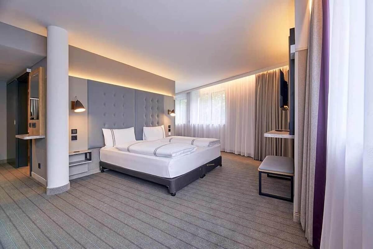 A modern hotel room with a large bed, a bedside table, a chair, and a large window with curtains.