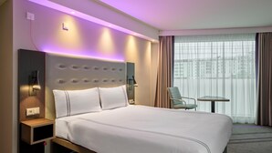 Hotelkamer met groot bed, nachtkastje, lamp, stoel en uitzicht op gebouwen door het raam.