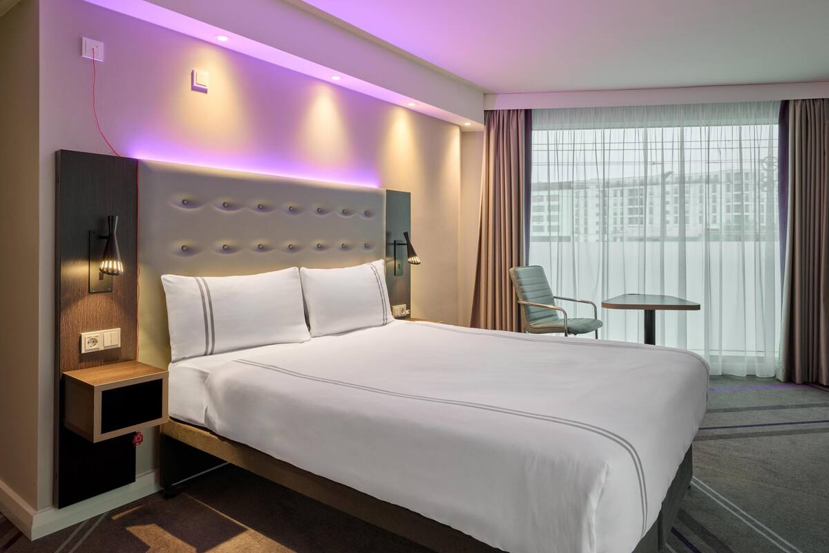 Una habitación de hotel con una cama grande, mesita de noche, lámpara, silla y vistas a edificios a través de la ventana.