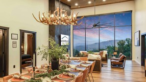 A modern dining area with a large table set for a meal, a chandelier, and a view of the mountains through large windows. - Incredible Mountain Views | Indoor Pool | Luxury (Sevierville)