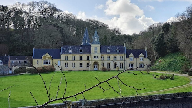 Un grand bâtiment historique doté de nombreuses fenêtres et d’une tour imposante, entouré d’une pelouse bien entretenue et d’un pont de pierre situé au premier plan.