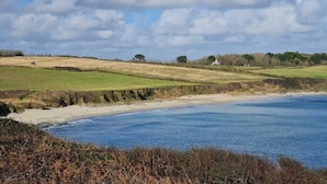 Un paysage côtier avec une plage de sable, des champs couverts d’herbe et une étendue d’eau sous un ciel partiellement nuageux.