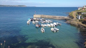 Un paysage côtier avec des bateaux amarrés à un quai, un phare à l’horizon et un petit bâtiment sur la plage.