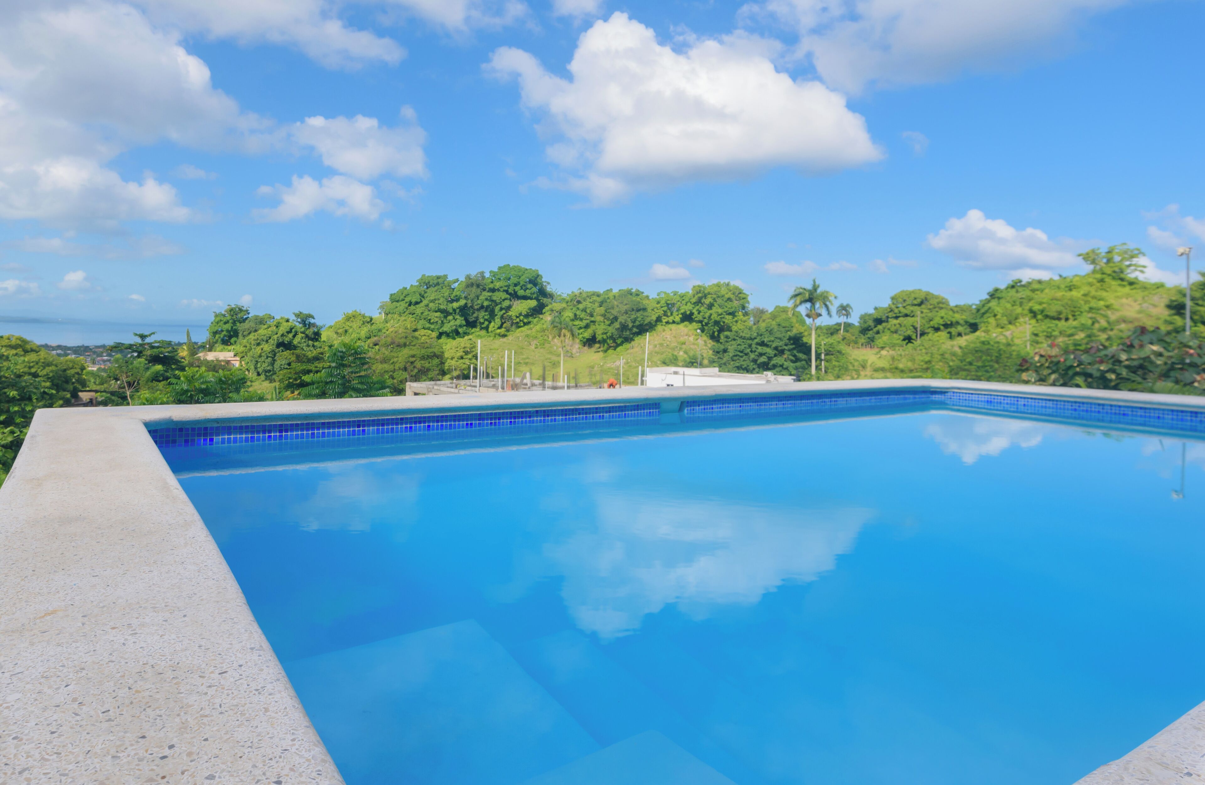 A clear blue swimming pool with a tiled edge, surrounded by lush greenery and a clear sky.