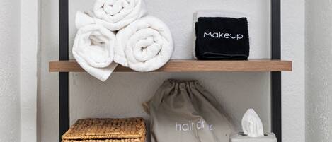 En hylla i badrummet med handdukar, en sminkväska, en väska för hårvårdsprodukter och en växt.