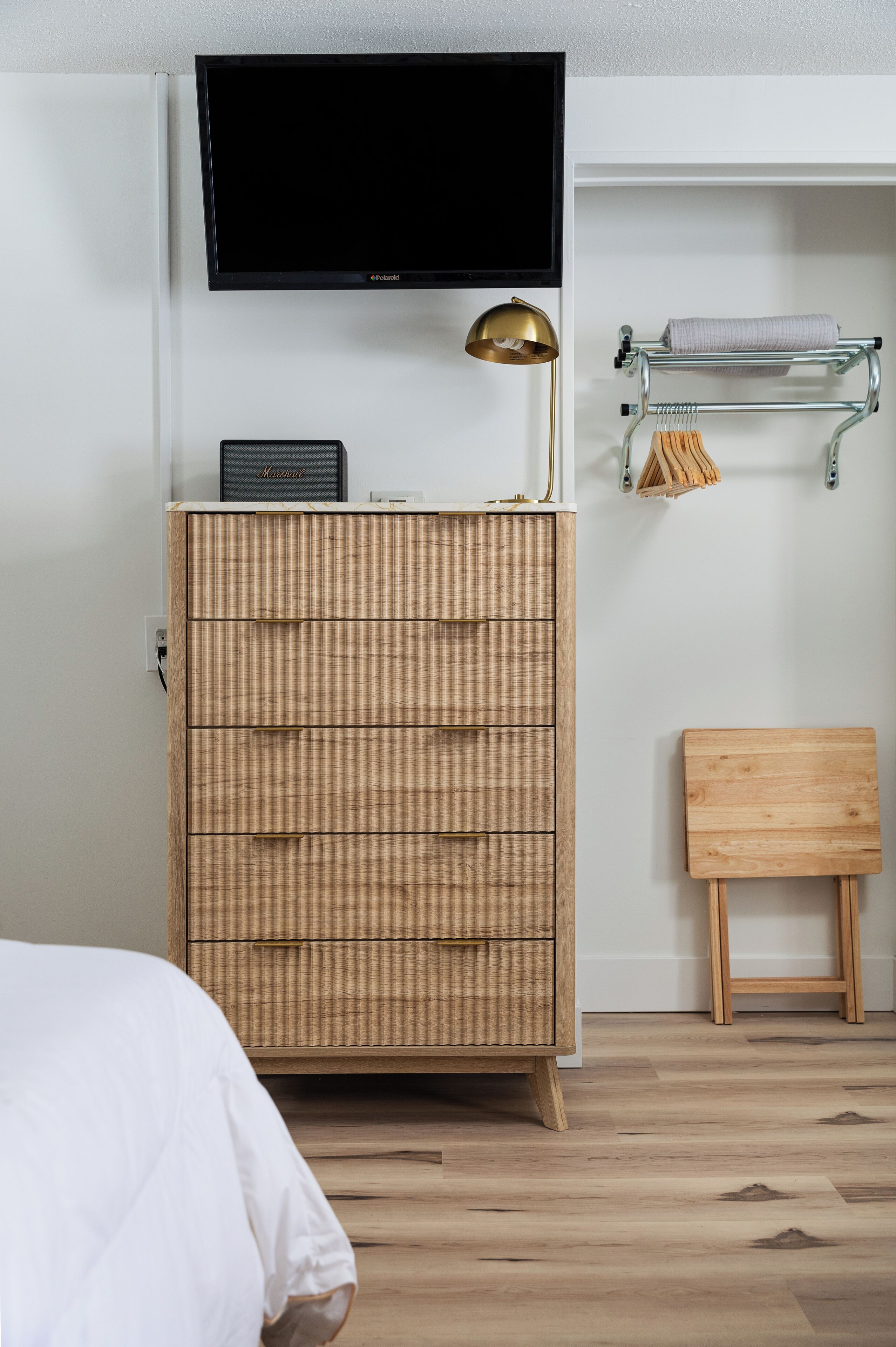A bedroom with a wooden dresser, a flat-screen TV, a lamp, a towel rack, and a wooden bench.