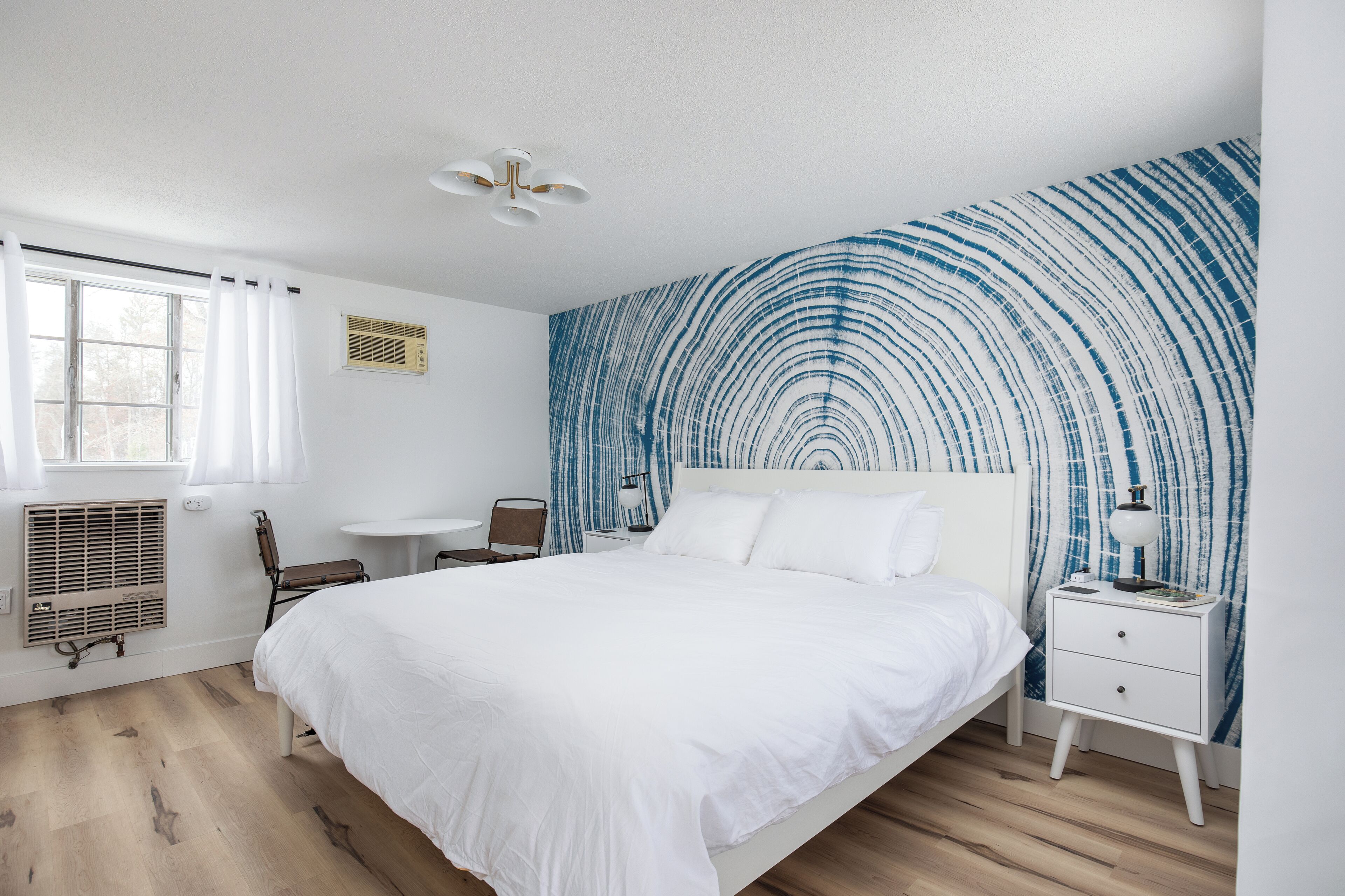 A bedroom with a bed, a bedside table, a chair, a window with curtains, a radiator, and a wall with a blue abstract pattern.