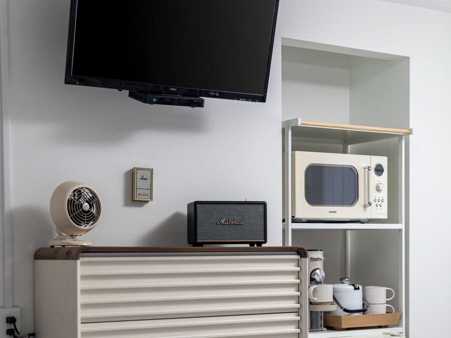 A room with a television mounted on the wall, a vintage-style microwave, a small fan, and a cabinet with drawers.