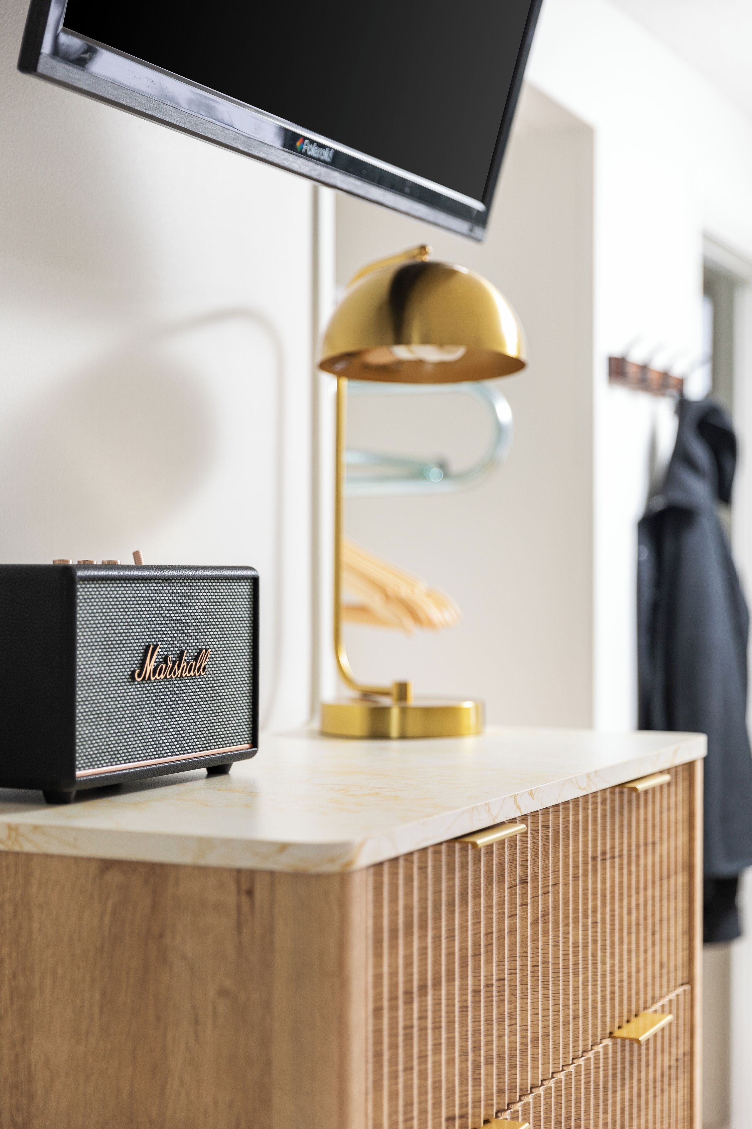 Una habitación moderna con un armario de madera, un altavoz Marshall, una lámpara dorada y un televisor montado en la pared.