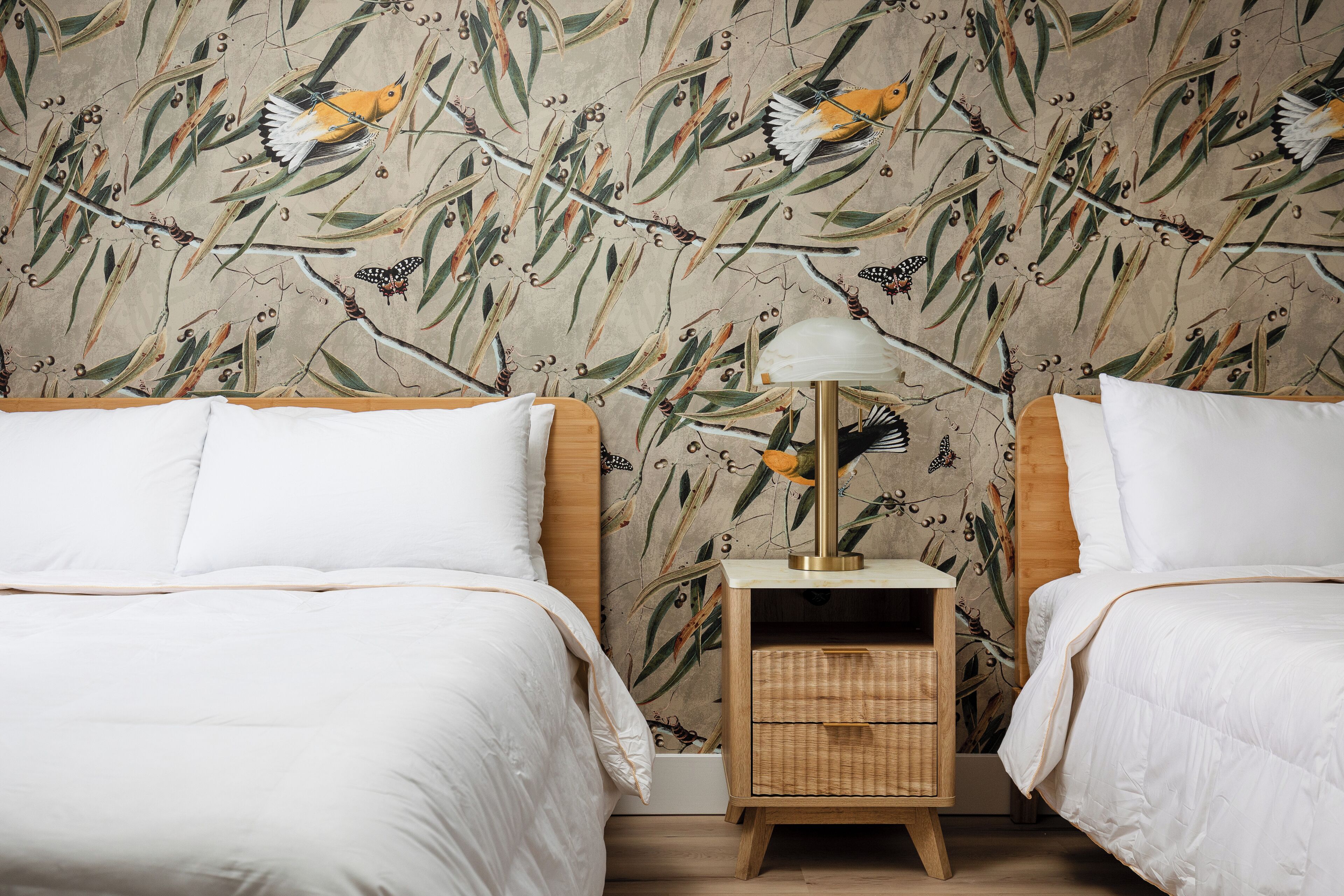 A bedroom with two beds, a nightstand with a lamp, and a wall with a nature-themed wallpaper.