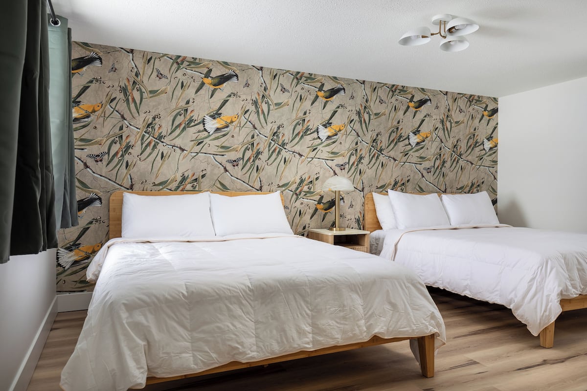 A room with two beds, a wooden floor, a wall with a nature-themed wallpaper, and a ceiling fan.