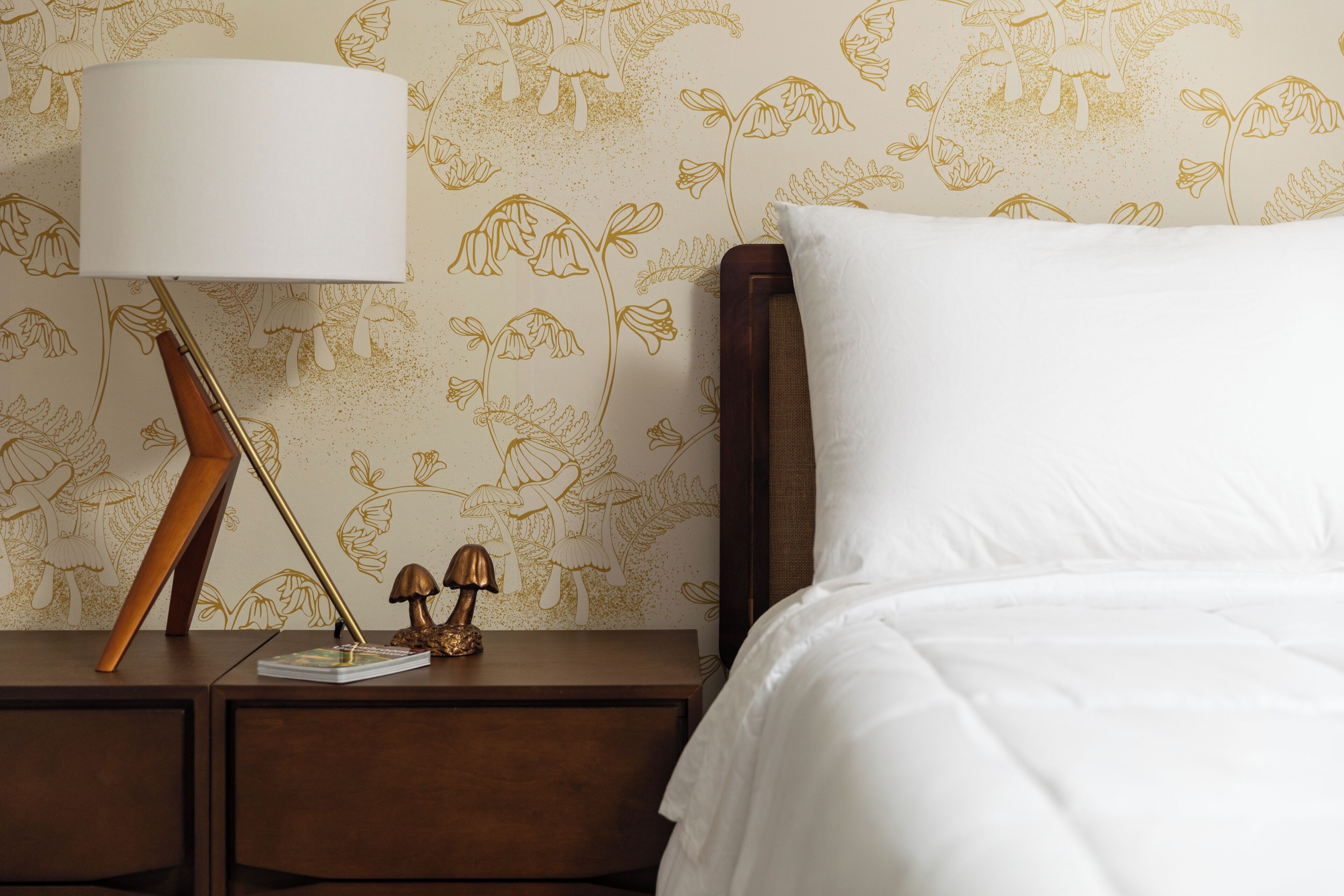 Un lit bien fait, avec un oreiller blanc et une tête de lit en bois, une table de chevet avec une lampe, et un mur orné d’un motif floral.
