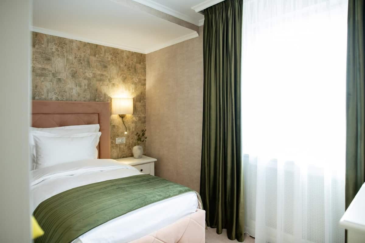 A hotel room with a bed, a nightstand, a lamp, and a view of the city through a window with curtains.
