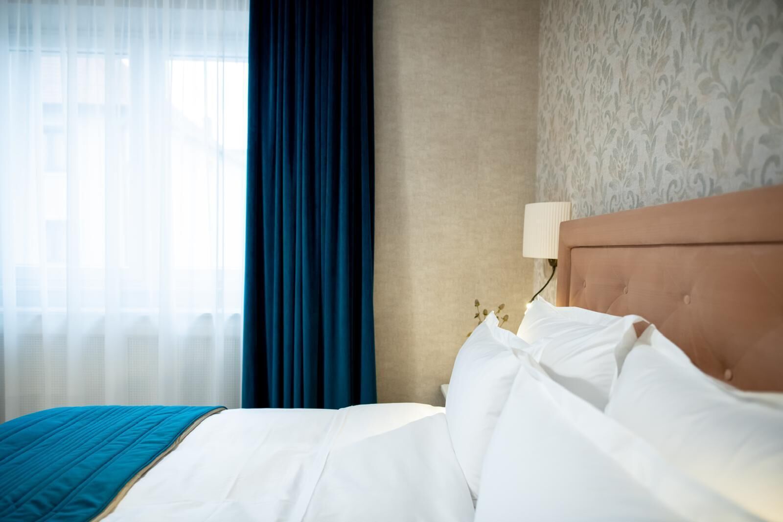 A hotel room with a bed, a window with sheer curtains, a dark blue curtain, a patterned wall, and a bedside lamp.