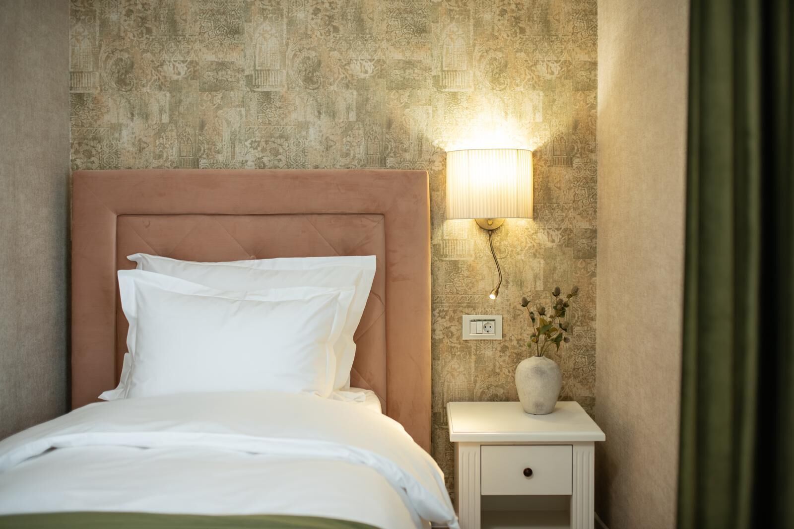 Uma cama bem arrumada com roupa de cama branca, cabeceira, mesa de cabeceira com abajur e um vaso com flores.