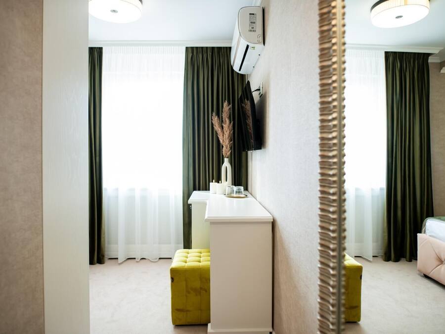 A modern hotel room with a white vanity, a yellow ottoman, a pink sofa, and a large window with green curtains.