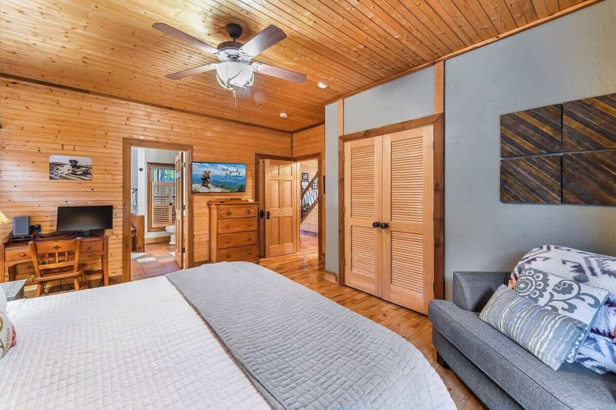 A wooden-floored room with a bed, a sofa, a desk with a computer, and a television. There is a door leading to another room and a ceiling fan.