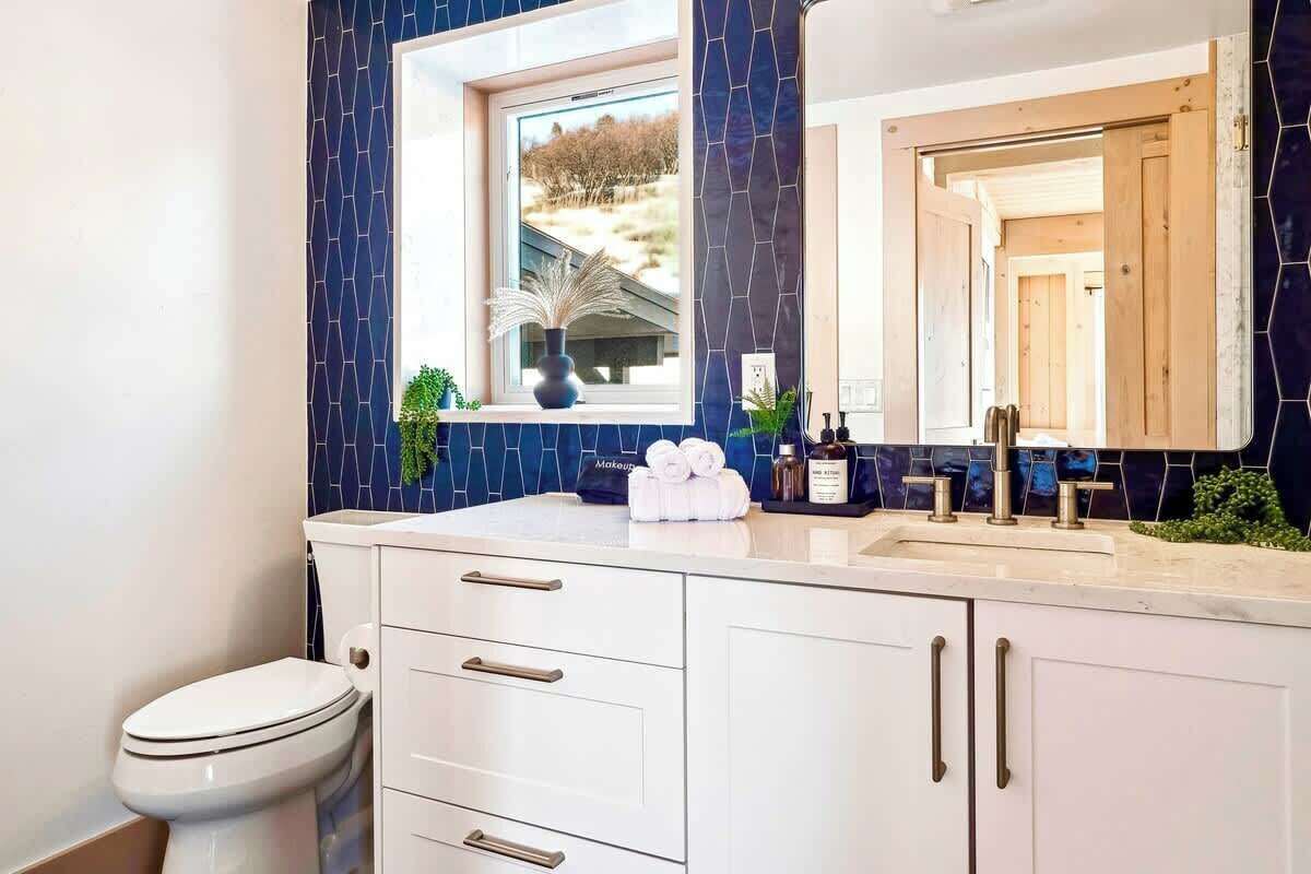 A bathroom with a white toilet, a sink with a mirror above it, and a window with a view of a house.