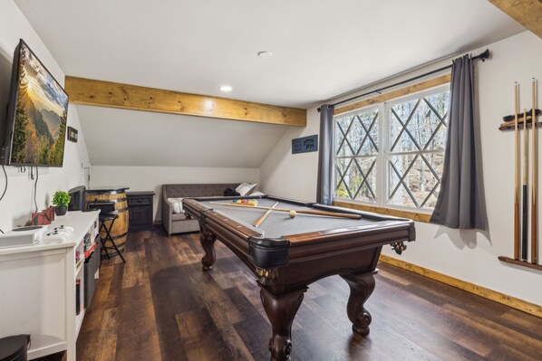 A pool table in a room with wooden flooring, a window with cross-bracing, and a television mounted on the wall. - High Top Cabin 3 Bedroom Cabin (Sevierville)