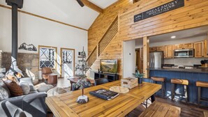 Een gezellig chaletinterieur met een houten eettafel, een open haard, een keuken met houten kasten en een trap naar de bovenverdieping.