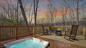 Een houten terras met een jacuzzi, buitenmeubilair en uitzicht op bomen en een zonsondergang.
