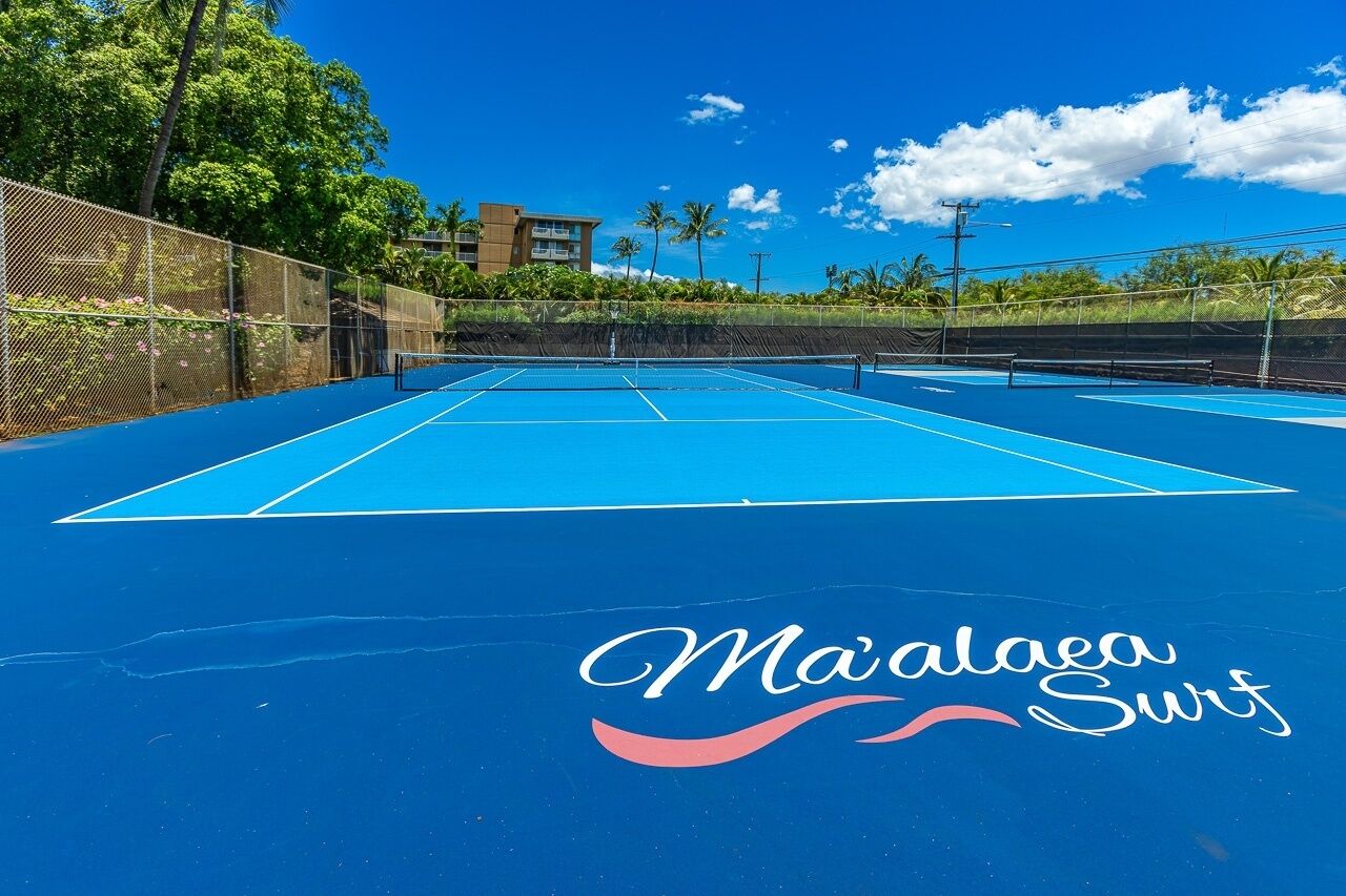 An outdoor tennis court with a blue surface and a net, surrounded by a fence and palm trees.