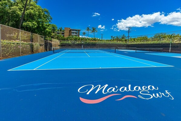 Un court de tennis extérieur doté d’une surface bleue et d’un filet, entouré d’une clôture et de palmiers.