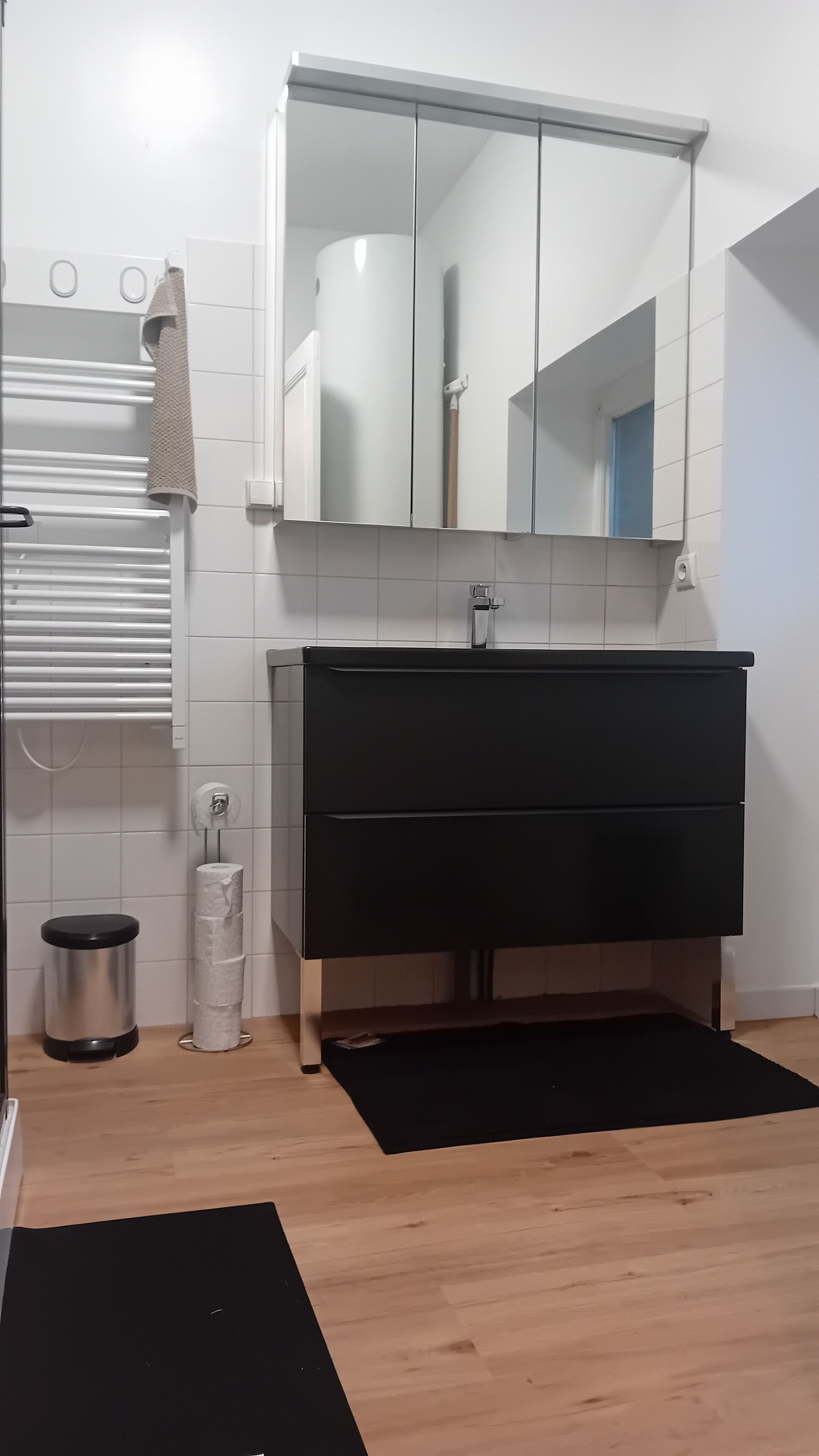 A bathroom with a black vanity, white tiles, a mirror, a towel rack, a toilet paper holder, and a trash bin.