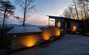 A modern cabin with a wooden exterior and a metal roof, situated in a forested area with a view of mountains in the background. - Mountain-Modern couples retreat in Bryson City with hot tub, fireplace, king bed free train tix (Bryson City)