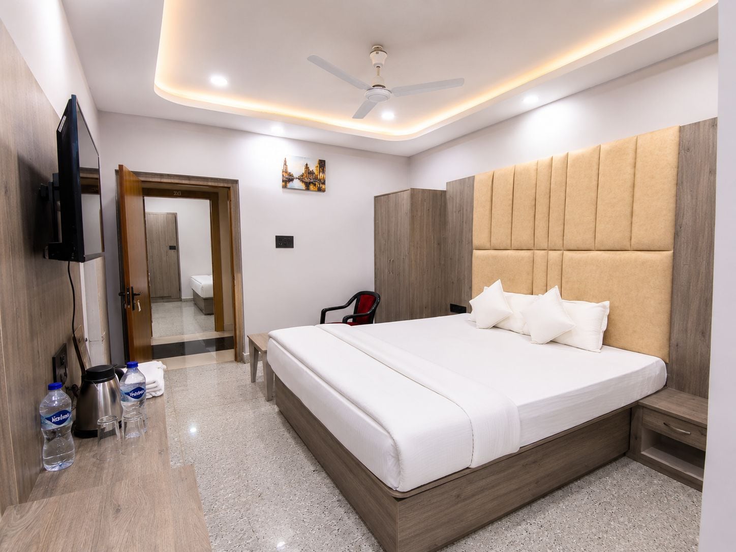 A hotel room with a large bed, a flat-screen TV mounted on the wall, a ceiling fan, and a wooden headboard.