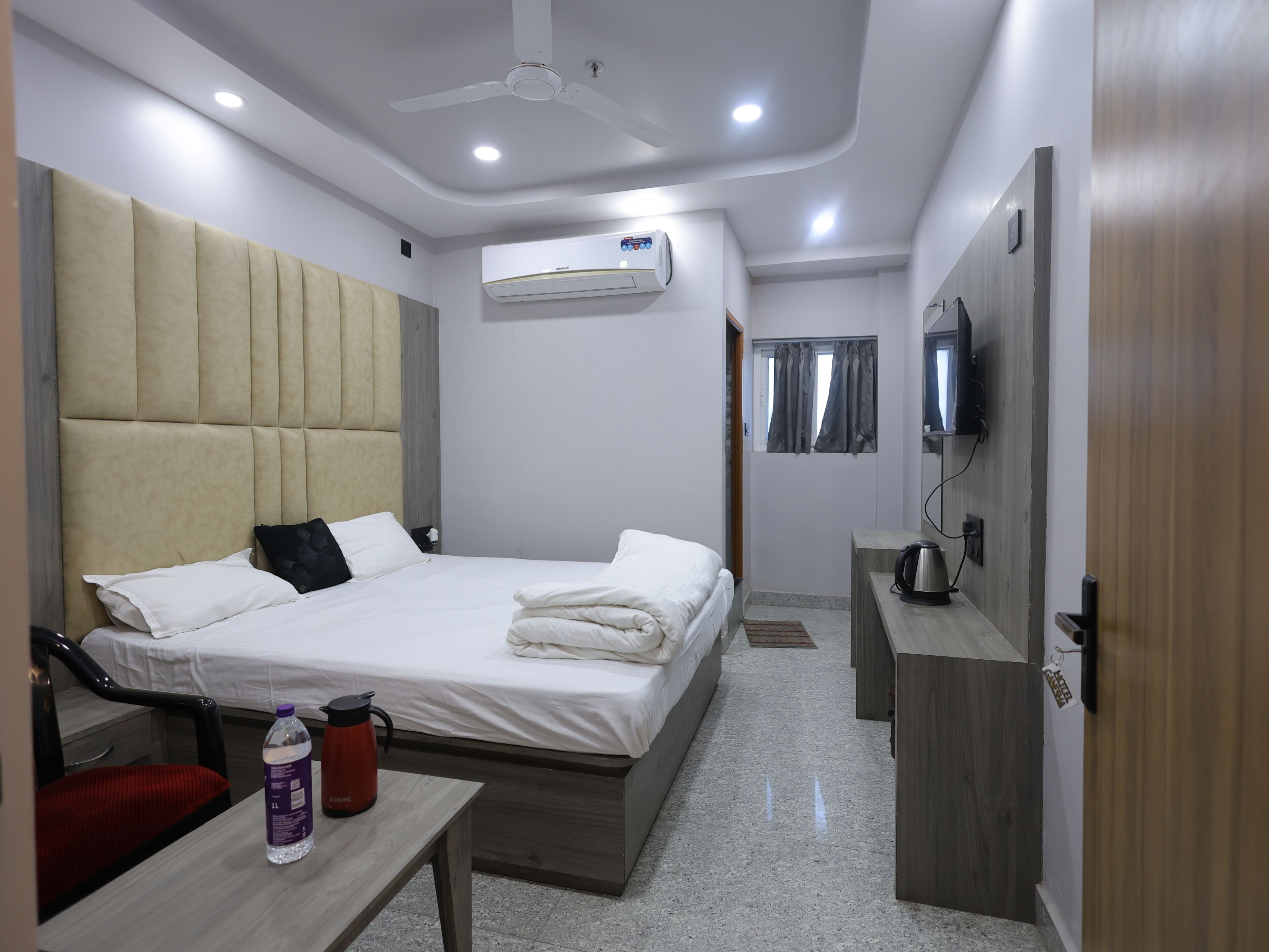 A hotel room with a bed, a table with a water bottle, a TV, and an air conditioning unit.