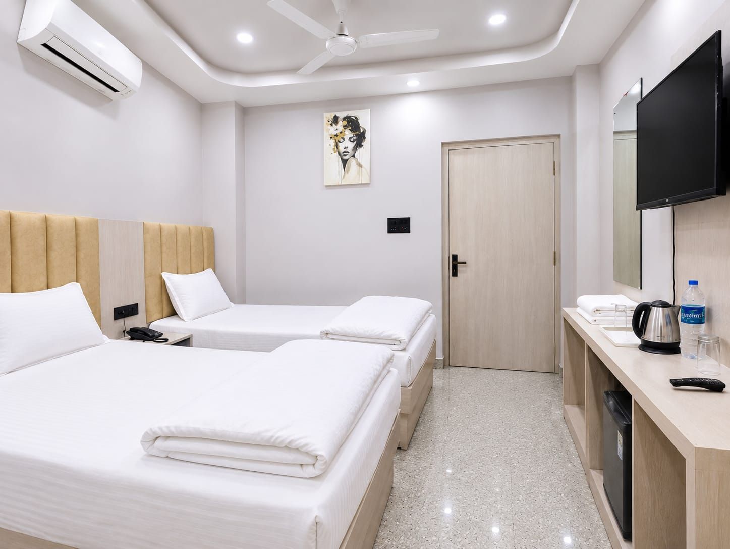 A hotel room with two beds, a ceiling fan, an air conditioner, a painting, a door, a minibar, and a television.