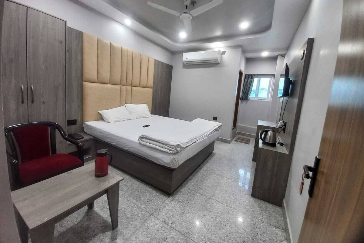 A modern hotel room with a bed, a desk, a chair, a wardrobe, an air conditioning unit, and a television.