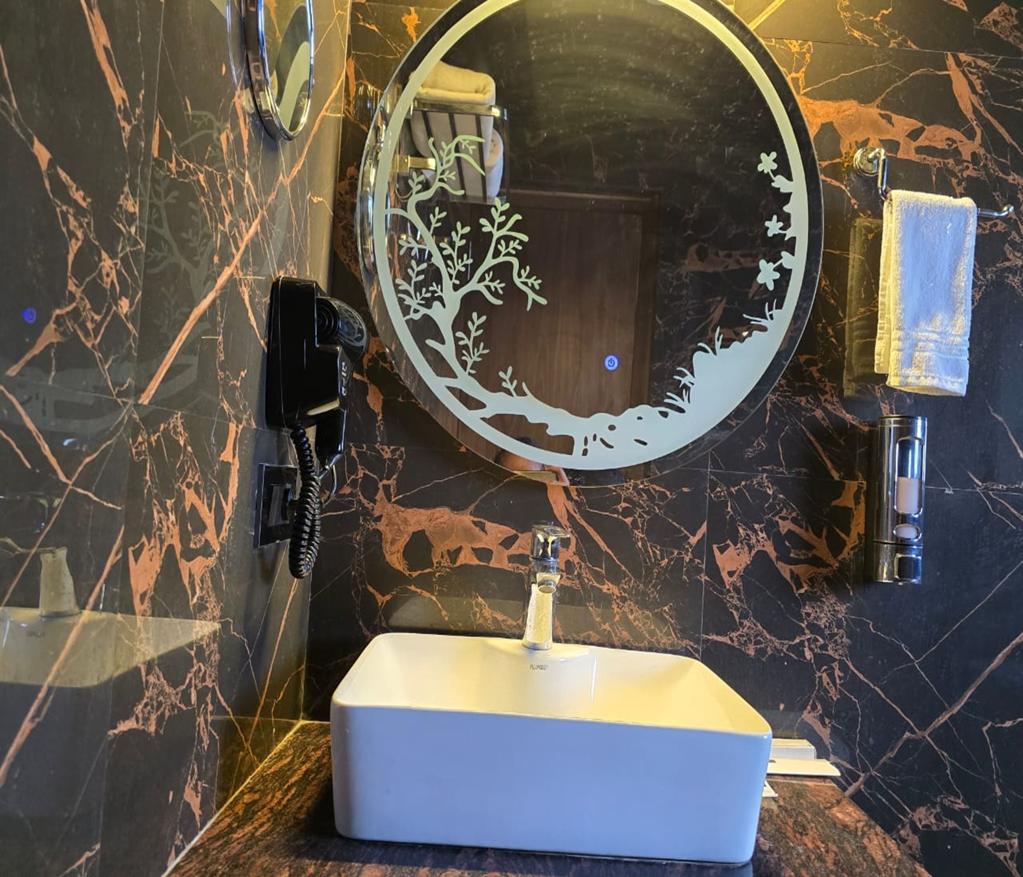Un baño con pared de mármol, un espejo redondo con diseño de árbol, un lavabo blanco y un perchero para toallas.