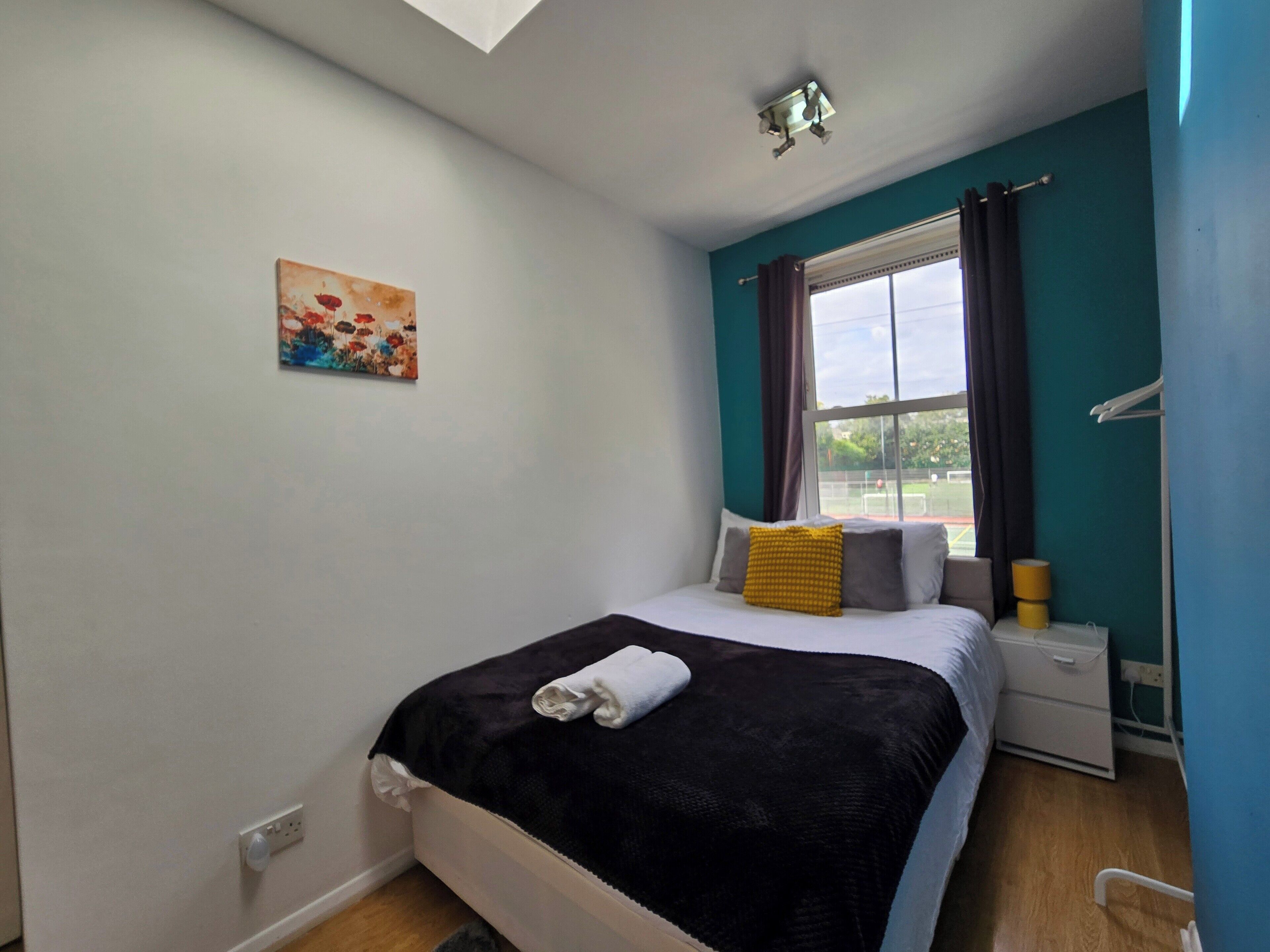 A bedroom with a bed, a nightstand, a window with curtains, a painting on the wall, and a ceiling light.