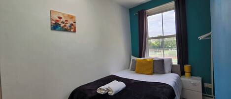 A bedroom with a bed, a nightstand, a window with curtains, a painting on the wall, and a ceiling light.