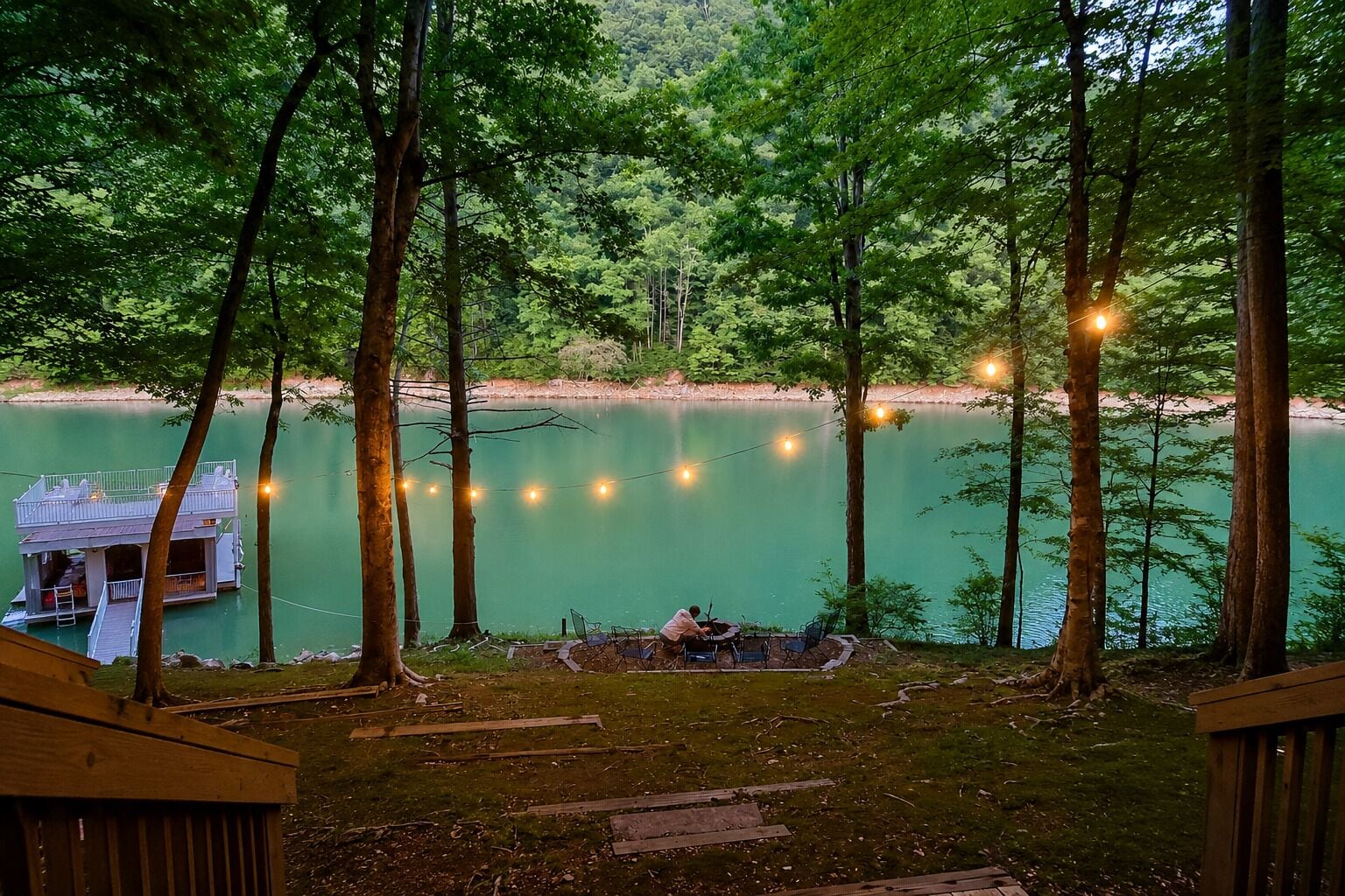 A lakeside deck with string lights, a dock, and a view of the lake surrounded by trees.