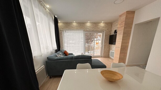 Un salon moderne avec un canapé gris, une table à manger blanche surmontée d’un saladier en fibres naturelles et une grande fenêtre agrémentée de rideaux légers.