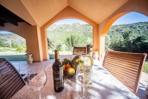 Mesa posta com garrafas de vinho e copos, uma cesta de frutas e vista para uma paisagem montanhosa através de uma janela em arco.
