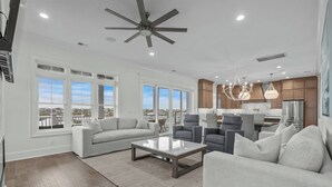 A spacious living room with a large gray sofa, a coffee table, and gray armchairs. There are large windows offering a view of the outside. - Tyson’s Corner: Luxe Marshfront Retreat w/ Pool (Murrells Inlet)