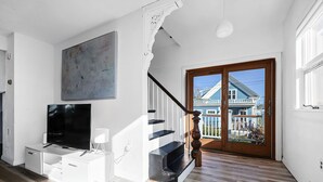 A spacious living room with a staircase leading to a balcony, a large painting on the wall, and a television on a white cabinet. - Painter's Perch: Rocky Neck (Gloucester)