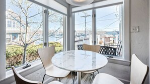 A room with a round table and four chairs, large windows with a view of buildings and trees, and a ceiling light fixture. - Painter's Perch: Rocky Neck (Gloucester)