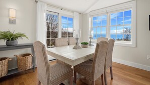 A dining area with a white table, wicker chairs, and a view of the sea through large windows. - Portside Overlook: Downtown Rockport Penthouse with stunning views (Rockport)