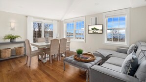A spacious living room with a grey sofa, a dining area with wicker chairs, and large windows offering a view of the outdoors. - Portside Overlook: Downtown Rockport Penthouse with stunning views (Rockport)
