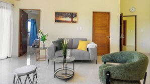 Una sala de estar con un sofá gris, una mesa de centro redonda de vidrio, un sillón verde y un cuadro de un árbol en la pared.