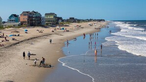 Una playa con casas al fondo, gente en la arena y olas acercándose.